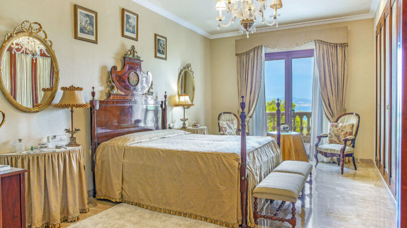 Elegantes Schlafzimmer der Luxusvilla mit antikem Himmelbett, Kronleuchter und Meerblick durch Terrassentür.