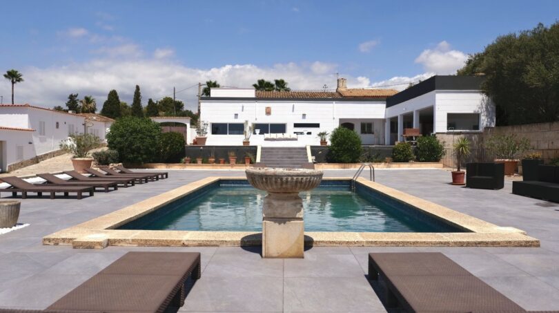 Außenbereich der Villa in Marratxí mit rechteckigem Pool, Steinbrunnen, Sonnenliegen und weißer Fassade.