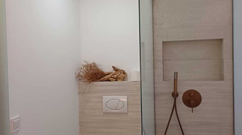Modernes Badezimmer mit Dusche, Regenduschkopf in Bronze, Wandhänge-WC und hellen Natursteinoptik-Fliesen.