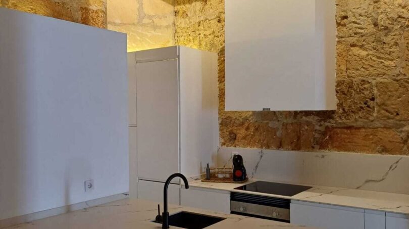 Moderne weiße Küche mit Marmorinsel, schwarzem Wasserhahn und rustikaler Steinwand in Erdgeschosswohnung Palma
