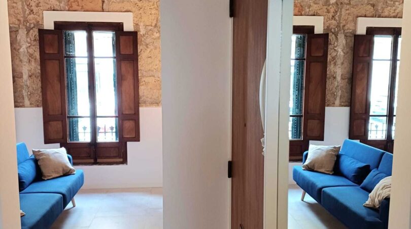 Flur der Erdgeschosswohnung in Palma mit Blick ins Wohnzimmer, blauem Sofa, Natursteinwand und Holzfensterläden