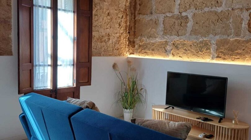 Wohnzimmer mit blauem Sofa, Natursteinwand, Holzbalkendecke und TV in Erdgeschosswohnung in Palma