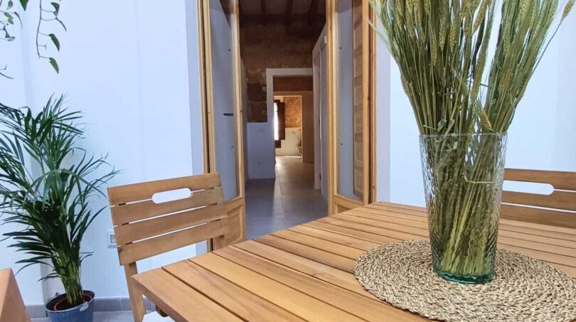 Überdachte Terrasse mit Holztisch und -stühlen, Grünpflanzen und Blick in den Innenbereich der Erdgeschosswohnung in Palma