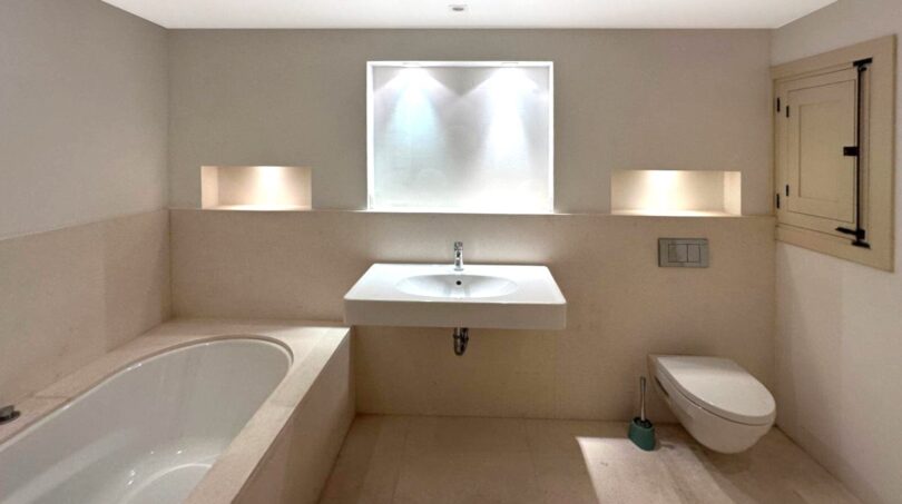 Modernes Badezimmer mit Einbaubadewanne, Wandwaschbecken, WC und eleganter Nischenbeleuchtung in Beige