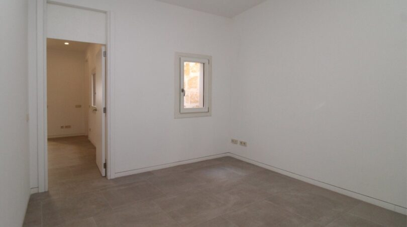 Dormitorio luminoso y vacío con piso de baldosas, pequeña ventana y paso al pasillo en el ático Born Palma