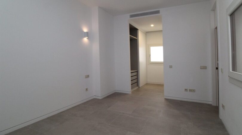 Dormitorio luminoso y vacío con piso de baldosas, aplique de pared y vista del vestidor incorporado en el ático Palma
