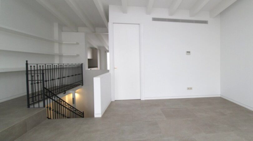 Planta superior luminosa del ático maisonette con escalera de hierro forjado, piso de hormigón y vigas de techo blancas