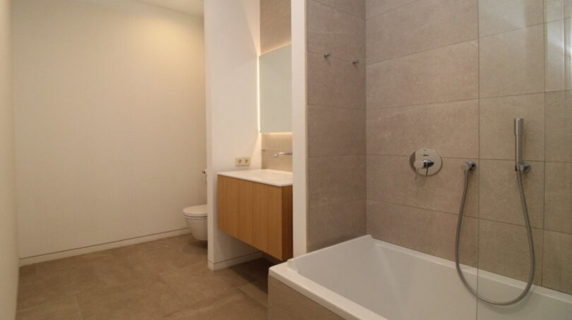 Baño moderno con bañera, tocador de madera, espejo iluminado y baldosas beige en el ático Palma