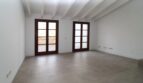 Espacio de estar luminoso y vacío con techo de vigas de madera, dos puertas francesas al balcón y piso de baldosas claro