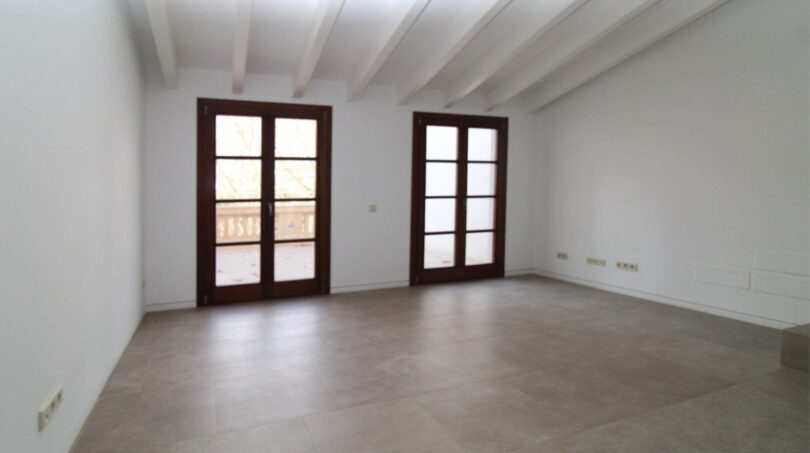 Espacio de estar luminoso y vacío con techo de vigas de madera, dos puertas francesas al balcón y piso de baldosas claro