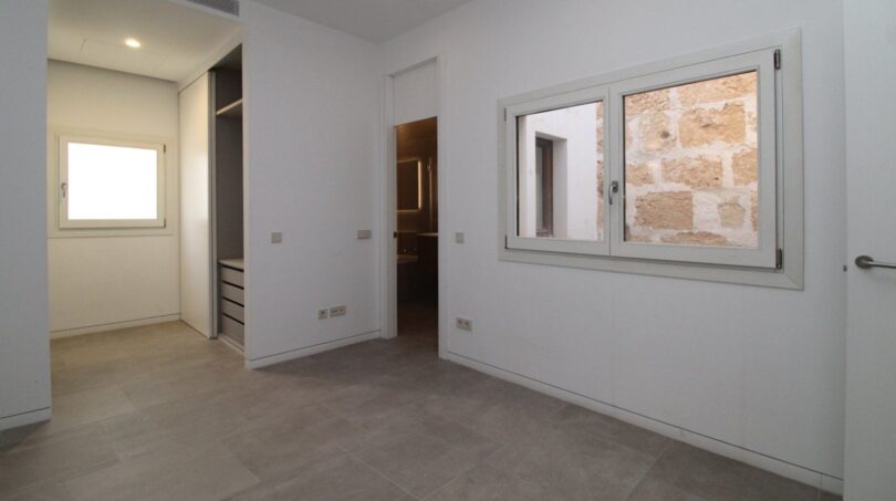 Dormitorio vacío con armario empotrado, luz natural y vista del muro de piedra histórico en el ático Borne Palma