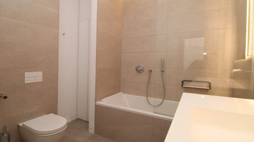 Baño moderno con bañera, mampara de vidrio, inodoro suspendido y baldosas beige en el ático Palma