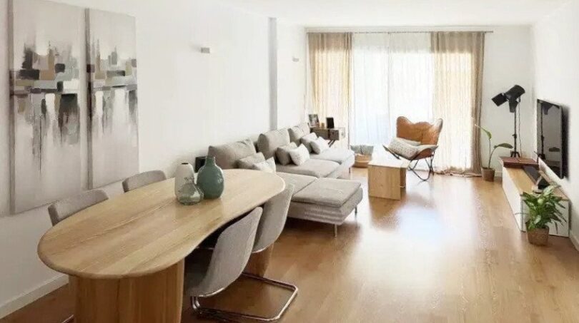 Sala de estar-comedor luminosa con suelo de madera, mesa de comedor ovalada, sofá gris y gran fachada de ventanas en Palma de Mallorca