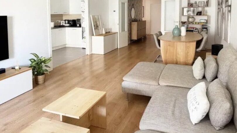 Sala de estar luminosa con sofá gris, suelo de madera y planta abierta con cocina y comedor en Palma