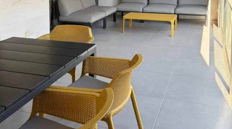 Balcón cubierto con sillas de ratán amarillas, mesa de comedor negra y grupo de sofás lounge gris en Palma de Mallorca.