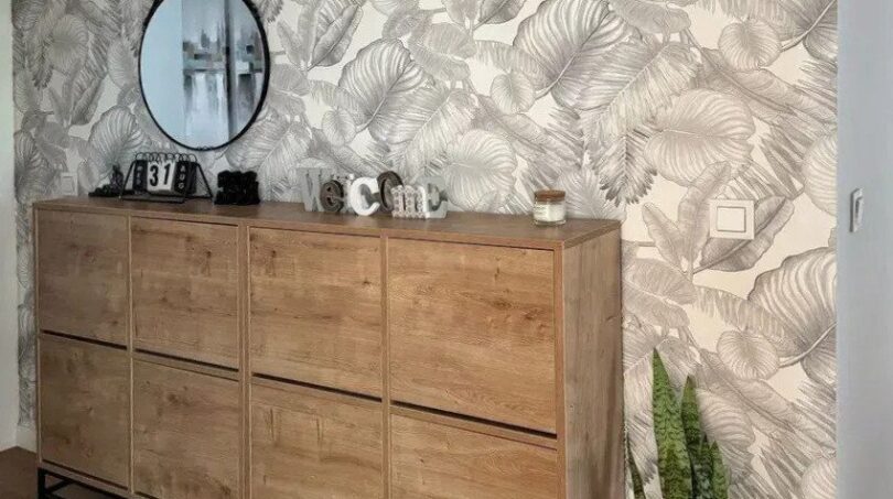 Zona de entrada con cómoda de madera, espejo redondo, Sansevieria y papel tapiz con patrón tropical en Palma de Mallorca