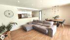 Helles Wohnzimmer mit grauem L-Sofa, Holzboden und offenem Durchgang zur Küche in modernem Einfamilienhaus Marratxí