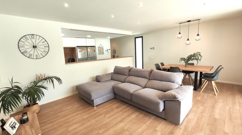 Helles Wohnzimmer mit grauem L-Sofa, Holzboden und offenem Durchgang zur Küche in modernem Einfamilienhaus Marratxí