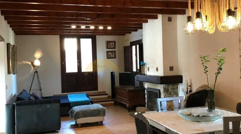 Rustikales Wohnzimmer der Finca Artà mit Holzbalkendecke, Kamin, blauer Couch und elegantem Esstisch.