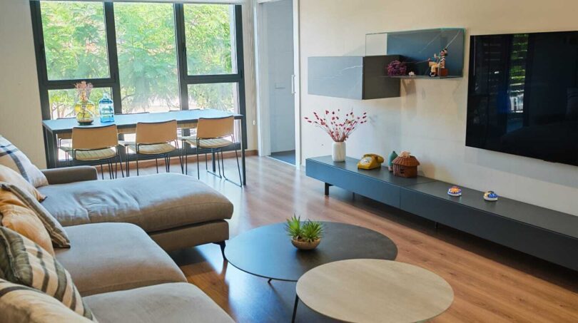 Helles Wohnzimmer mit grauem Sofa, Holzboden, TV-Wand und großen Fenstern mit Grünblick in Palma