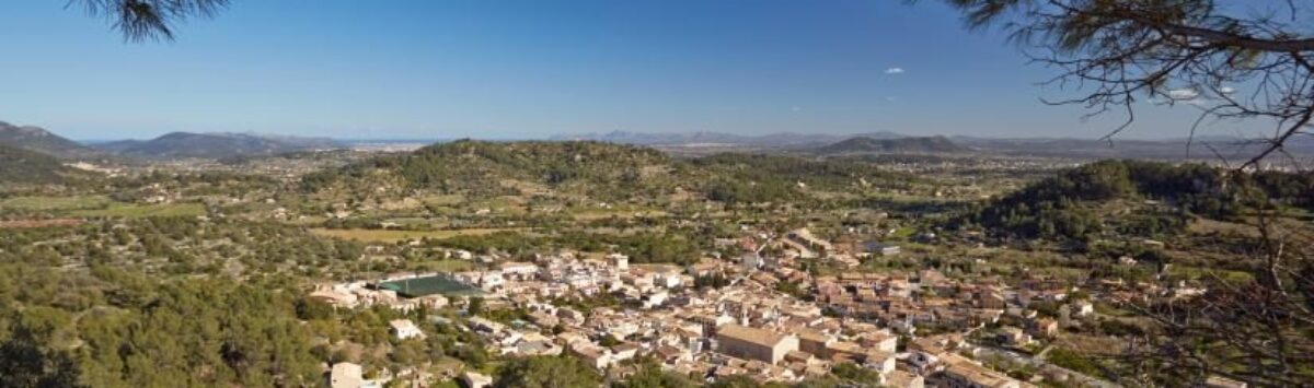 Vista panorámica de Mancor de la Vall con casas de piedra natural, rodeadas de colinas verdes y paisaje extenso.