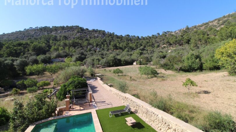 Finca Llucmajor: Pool mit Liegewiese, Natursteinmauer und Blick auf bewaldeten Hexenberg unter blauem Himmel.