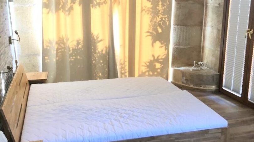 Schlafzimmer der Natur-Finca in Llucmajor mit Holzbett, Steinwänden und lichtdurchfluteten Vorhängen.