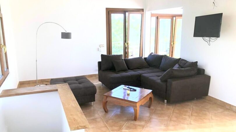 Helles Wohnzimmer der Finca in Llucmajor mit dunklem Ecksofa, Holzbalkendecke und Terrassentüren mit Naturblick.