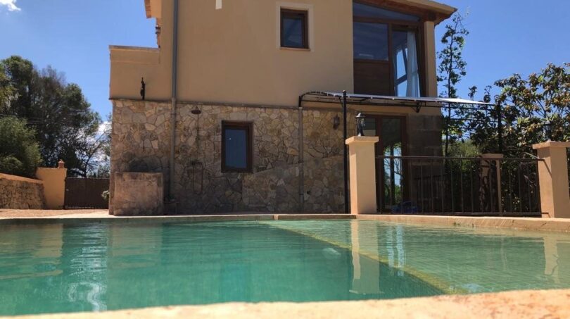 Finca Llucmajor: Außenansicht mit Pool, Natursteinwand und großzügiger Glasfront, eingebettet in mediterrane Landschaft.