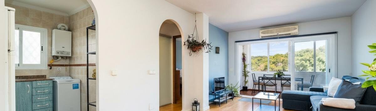 Helles Wohnzimmer mit Parkettboden, Klimaanlage, Balkon mit Naturblick und Einblick in Küche mit Waschmaschine.