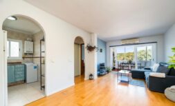 Helles Wohnzimmer mit Parkettboden, Klimaanlage, Balkon mit Naturblick und Einblick in Küche mit Waschmaschine.