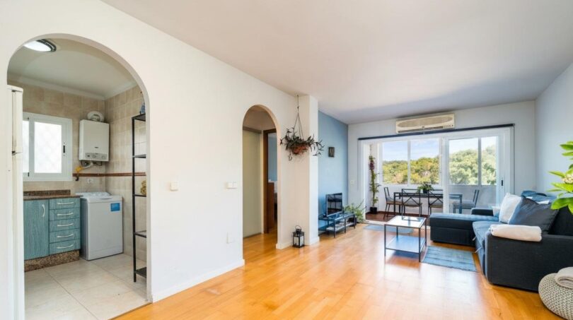 Helles Wohnzimmer mit Parkettboden, Klimaanlage, Balkon mit Naturblick und Einblick in Küche mit Waschmaschine.