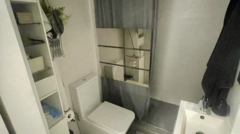 Baño compacto con inodoro, lavamanos, ducha con cortina gris y unidad de estantería en apartamento del casco antiguo de Palma.