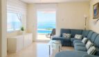 Helles Wohnzimmer der First-Line-Villa in Llucmajor mit blauem Ecksofa, Marmorboden und direktem Meerblick durch Glastür.
