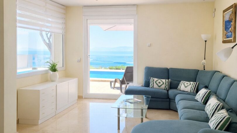 Helles Wohnzimmer der First-Line-Villa in Llucmajor mit blauem Ecksofa, Marmorboden und direktem Meerblick durch Glastür.