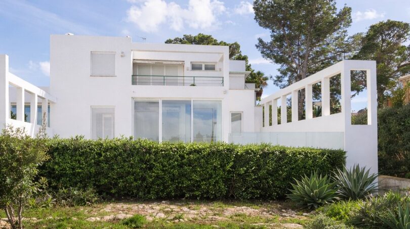 Außenansicht der weißen First-Line-Villa in Llucmajor mit Balkon, Glasfront und gepflegter Gartenanlage.