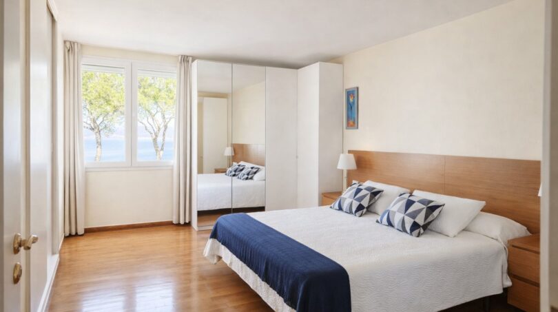 Helles Schlafzimmer der First-Line-Villa in Llucmajor mit Doppelbett, Holzboden und Meerblick durchs Fenster.