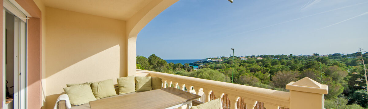 Sonnige Terrasse des Reihenhauses in Cales de Mallorca mit Sitzecke, Holztisch und Meerblick über grüne Pinien.
