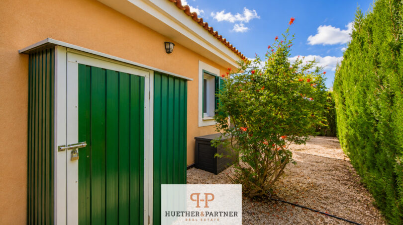Gartenbereich des Luxus-Chalets in Cala Murada mit grünem Metallschuppen, blühendem Strauch und Zypressenhecke.