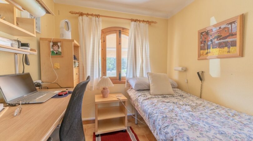 Helles Kinderzimmer der Finca Campanet mit Einzelbett, Schreibtisch, Laptop und Rundbogenfenster mit Gardinenschmuck.