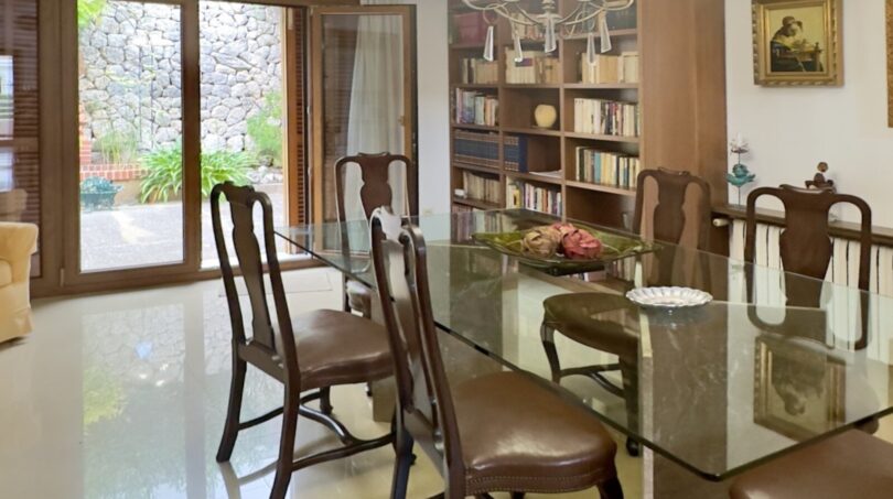 Elegante comedor con mesa de cristal, sillas de madera, lámpara de araña y estantería, con vista al jardín en Palma