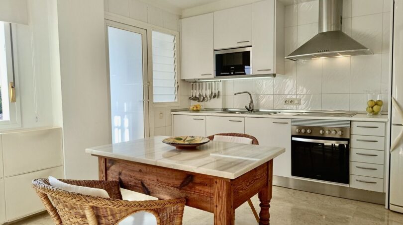 Bright kitchen of the Mallorca apartment with white cabinets, stainless steel hood and rustic wooden table with wicker chairs.