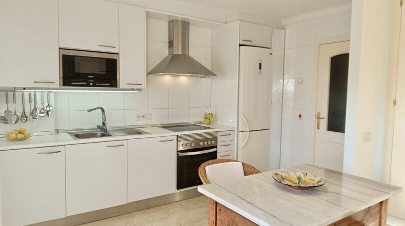 Bright built-in kitchen with white fronts, stainless steel range hood, oven and rustic marble dining table in Mallorca apartment.