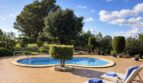 Finca Campanet: Ovaler Pool mit Terrasse, Liegestühlen und üppigem Garten unter blauem Himmel.