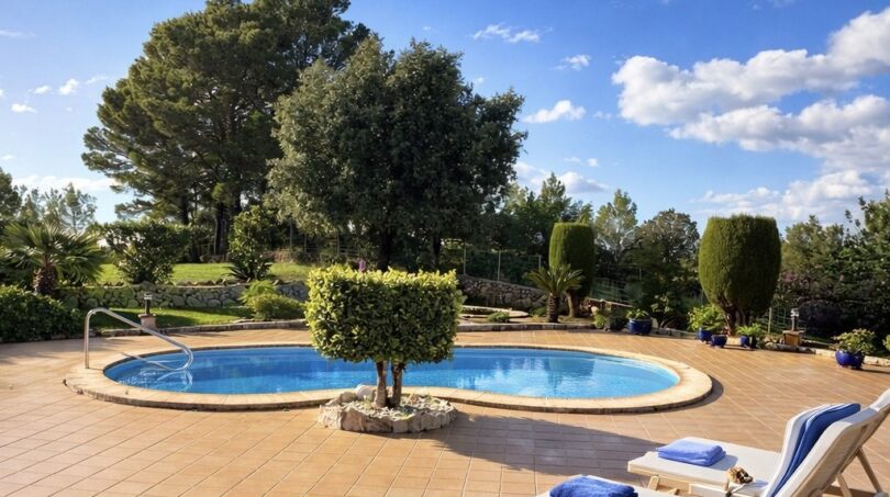 Finca Campanet: Ovaler Pool mit Terrasse, Liegestühlen und üppigem Garten unter blauem Himmel.