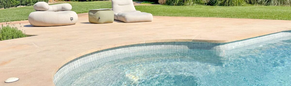 Curved pool with mosaic tiles, beige terrace and lounge furniture, surrounded by lush garden of the stone finca Felanitx