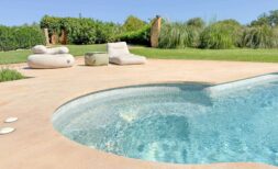 Piscina curva con azulejos de mosaico, terraza beige y muebles de lounge, rodeada por jardín verde de la finca de piedra Felanitx