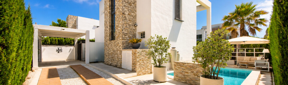 Außenansicht der Luxus-Bauhausvilla in Cala Murada mit Pool, Natursteinturm, weißer Fassade und gepflegtem Garten.