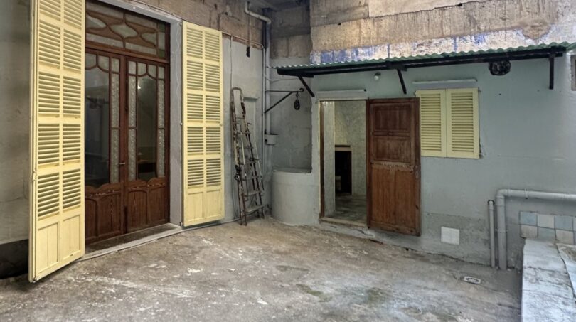 Innenhof des Stadthauses in Manacor mit gelben Holzfensterläden, Betonboden und Holztüren, renovierungsbedürftig.