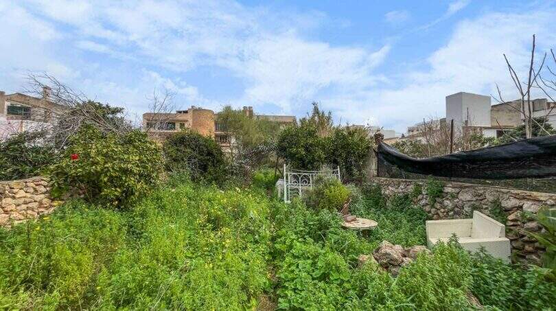 An overgrown garden with lush vegetation, stone walls, and white garden furniture, surrounded by houses in Felanitx.